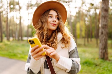 Telefonlu ve şapkalı genç bir kadın güneşli bir parkta eğleniyor. Güzel bir kadın gün batımında yürümekten ve mesajlaşmaktan zevk alıyor. Blog yazma, boş zaman konsepti. Etkin yaşam biçimi.
