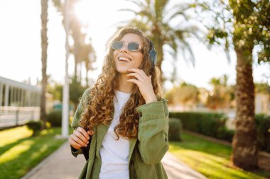 Güneş gözlüklü ve kablosuz kulaklıklı neşeli kadın palmiye ağaçlarıyla çevrili güneşli bir günün tadını çıkarıyor. Güzel bir turist müzik dinler ve gün batımında eğlenir. Yürüme, teknoloji konsepti.