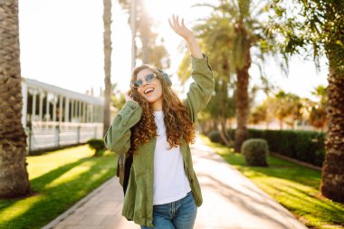 Güneş gözlüklü ve kablosuz kulaklıklı neşeli kadın palmiye ağaçlarıyla çevrili güneşli bir günün tadını çıkarıyor. Güzel bir turist müzik dinler ve gün batımında eğlenir. Yürüme, teknoloji konsepti.