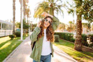 Güneş gözlüklü ve kablosuz kulaklıklı neşeli kadın palmiye ağaçlarıyla çevrili güneşli bir günün tadını çıkarıyor. Güzel bir turist müzik dinler ve gün batımında eğlenir. Yürüme, teknoloji konsepti.