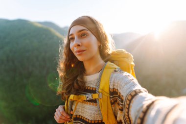 Gün batımında dağ yamacında selfie çeken genç bir gezginin portresi. Selfie zamanı. Sırt çantalı güzel bir kadın dağ doğasını ve dışarıda blog yazmayı seviyor. Seyahat, günlük tutma kavramı.