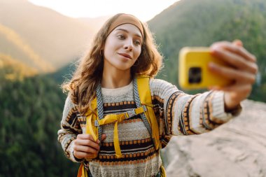 Gün batımında dağ yamacında selfie çeken genç bir gezginin portresi. Selfie zamanı. Sırt çantalı güzel bir kadın dağ doğasını ve dışarıda blog yazmayı seviyor. Seyahat, günlük tutma kavramı.