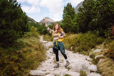 Güneşli bir günde dağ yolunda sarı bir sırt çantasıyla yürüyüş yapan bir bayan yürüyüşçü. Güzel bir kadın dağ manzarasının tadını çıkarıyor. Macera konsepti, yürüyüş. Etkin yaşam biçimi.
