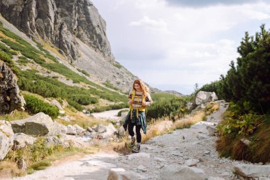 Güneşli bir günde dağ yolunda sarı bir sırt çantasıyla yürüyüş yapan bir bayan yürüyüşçü. Güzel bir kadın dağ manzarasının tadını çıkarıyor. Macera konsepti, yürüyüş. Etkin yaşam biçimi.