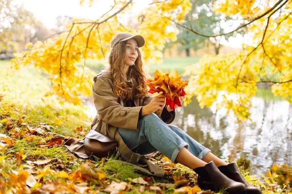 Mutlu kadın güneşli bir sonbahar parkında bir buket parlak yaprakla eğleniyor. Günbatımının tadını çıkaran günlük giysiler içinde güzel bir turist. Yürüme konsepti. Sonbahar atmosferi.