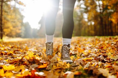 Yürüyüş botlarıyla, renkli sonbahar yapraklarıyla çevrili yürüyüş yolunda duran bir yürüyüşçünün bacaklarının yakın çekimi. Yağmurluk ve çizmeli bir kadın parkta sonbahar gününün tadını çıkarır. Macera konsepti.
