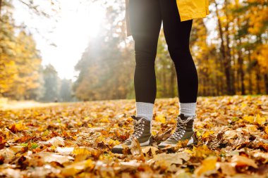 Yürüyüş botlarıyla, renkli sonbahar yapraklarıyla çevrili yürüyüş yolunda duran bir yürüyüşçünün bacaklarının yakın çekimi. Yağmurluk ve çizmeli bir kadın parkta sonbahar gününün tadını çıkarır. Macera konsepti.