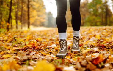 Yürüyüş botlarıyla, renkli sonbahar yapraklarıyla çevrili yürüyüş yolunda duran bir yürüyüşçünün bacaklarının yakın çekimi. Yağmurluk ve çizmeli bir kadın parkta sonbahar gününün tadını çıkarır. Macera konsepti.