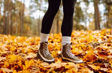 Yürüyüş botlarıyla, renkli sonbahar yapraklarıyla çevrili yürüyüş yolunda duran bir yürüyüşçünün bacaklarının yakın çekimi. Yağmurluk ve çizmeli bir kadın parkta sonbahar gününün tadını çıkarır. Macera konsepti.