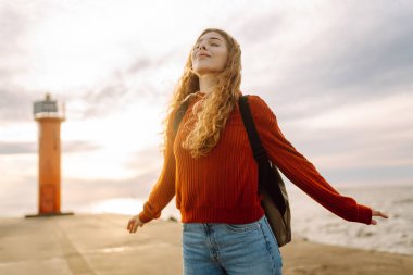 Sırt çantalı genç bir gezgin deniz feneri manzaralı iskele boyunca yürüyor. Gün batımında deniz kenarında özgür hisseden güzel bir kadın. Aktif yaşam tarzı. Doğa ve seyahat kavramı.