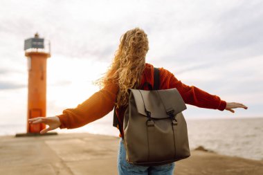 Sırt çantalı genç bir gezgin deniz feneri manzaralı iskele boyunca yürüyor. Gün batımında deniz kenarında özgür hisseden güzel bir kadın. Aktif yaşam tarzı. Doğa ve seyahat kavramı.