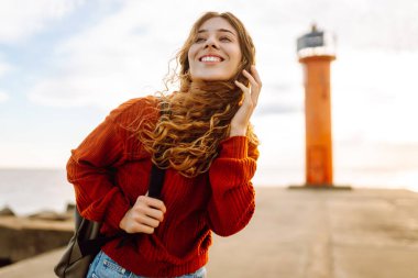 Sırt çantalı genç bir gezgin deniz feneri manzaralı iskele boyunca yürüyor. Gün batımında deniz kenarında özgür hisseden güzel bir kadın. Aktif yaşam tarzı. Doğa ve seyahat kavramı.