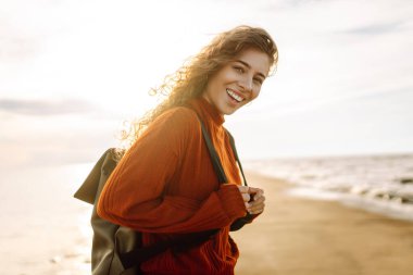 Sırt çantalı genç bir gezgin deniz feneri manzaralı iskele boyunca yürüyor. Gün batımında deniz kenarında özgür hisseden güzel bir kadın. Aktif yaşam tarzı. Doğa ve seyahat kavramı.