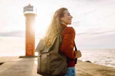 Sırt çantalı genç bir gezgin deniz feneri manzaralı iskele boyunca yürüyor. Gün batımında deniz kenarında özgür hisseden güzel bir kadın. Aktif yaşam tarzı. Doğa ve seyahat kavramı.