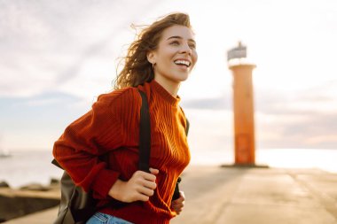 Sırt çantalı genç bir gezgin deniz feneri manzaralı iskele boyunca yürüyor. Gün batımında deniz kenarında özgür hisseden güzel bir kadın. Aktif yaşam tarzı. Doğa ve seyahat kavramı.