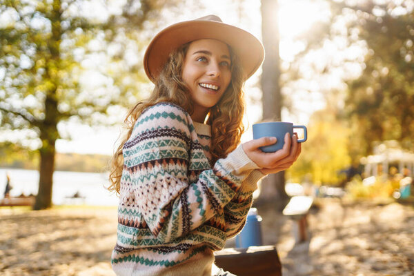 Portrait of woman in hat with cup of hot tea from thermos sitting on bench in autumn park. Beautiful female traveler enjoying hot drink on sunny day. Rest and relaxation concept.