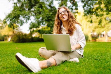 Gözlüklü ve bilgisayarlı mutlu kadın güneşli bir parkta yeşil çimlerin üzerinde oturuyor. Dışarıda dizüstü bilgisayarla çalışan genç bir bayan serbest yazar. Doğa kavramı, uzak çalışma ve teknoloji.