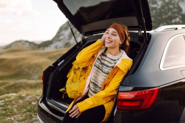 Sarı yağmurluklu ve sırt çantalı neşeli bir kadın arabanın bagajında oturuyor ve dağ manzarasının tadını çıkarıyor. Genç gezgin arabayla seyahat ederken özgürlüğü hisseder. Seyahat tutkusu. Ulaşım ve özgürlük kavramı.