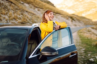 Sarı yağmurluklu ve sırt çantalı neşeli bir kadın arabanın bagajında oturuyor ve dağ manzarasının tadını çıkarıyor. Genç gezgin arabayla seyahat ederken özgürlüğü hisseder. Seyahat tutkusu. Ulaşım ve özgürlük kavramı.