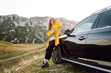 Sarı yağmurluklu ve sırt çantalı neşeli bir kadın arabanın bagajında oturuyor ve dağ manzarasının tadını çıkarıyor. Genç gezgin arabayla seyahat ederken özgürlüğü hisseder. Seyahat tutkusu. Ulaşım ve özgürlük kavramı.