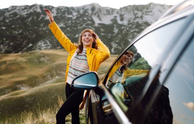 Sarı yağmurluklu ve sırt çantalı neşeli bir kadın arabanın bagajında oturuyor ve dağ manzarasının tadını çıkarıyor. Genç gezgin arabayla seyahat ederken özgürlüğü hisseder. Seyahat tutkusu. Ulaşım ve özgürlük kavramı.