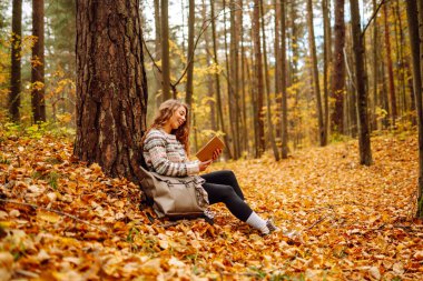 Elinde kitap olan rahat bir kadın sonbahar parkında bir ağacın yanında oturuyor. Şapkalı ve sırt çantalı güzel bir gezgin parlak yapraklarla çevrili bir kitap okuyor. Rahatlama konsepti, mevsimlik.
