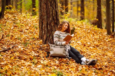 Elinde kitap olan rahat bir kadın sonbahar parkında bir ağacın yanında oturuyor. Şapkalı ve sırt çantalı güzel bir gezgin parlak yapraklarla çevrili bir kitap okuyor. Rahatlama konsepti, mevsimlik.