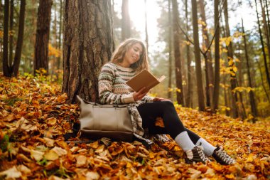 Elinde kitap olan rahat bir kadın sonbahar parkında bir ağacın yanında oturuyor. Şapkalı ve sırt çantalı güzel bir gezgin parlak yapraklarla çevrili bir kitap okuyor. Rahatlama konsepti, mevsimlik.