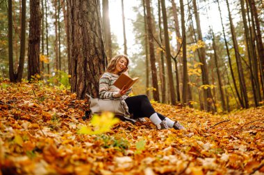 Elinde kitap olan rahat bir kadın sonbahar parkında bir ağacın yanında oturuyor. Şapkalı ve sırt çantalı güzel bir gezgin parlak yapraklarla çevrili bir kitap okuyor. Rahatlama konsepti, mevsimlik.