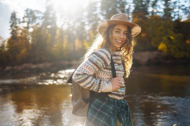 Nehir kenarında sırt çantalı mutlu bir kadın güneşli bir günün tadını çıkarıyor. Dişi gezgin kendini özgür hissediyor ve sonbahar ormanında suyun kenarında eğleniyor. Macera ve zevk konsepti.