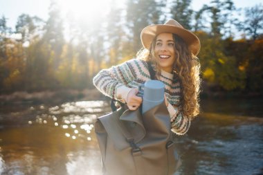 Nehir kenarında sırt çantalı mutlu bir kadın güneşli bir günün tadını çıkarıyor. Dişi gezgin kendini özgür hissediyor ve sonbahar ormanında suyun kenarında eğleniyor. Macera ve zevk konsepti.