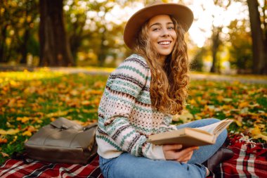 Sonbahar parkında kırmızı battaniyede oturan şapkalı genç bir kadın. Kitap okumayı seven mutlu bir kadın ve sarı yapraklı çayırda güneşli bir gün. Dinlenme kavramı, rahatlama..