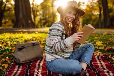 Sonbahar parkında kırmızı battaniyede oturan şapkalı genç bir kadın. Kitap okumayı seven mutlu bir kadın ve sarı yapraklı çayırda güneşli bir gün. Dinlenme kavramı, rahatlama..