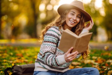 Sonbahar parkında kırmızı battaniyede oturan şapkalı genç bir kadın. Kitap okumayı seven mutlu bir kadın ve sarı yapraklı çayırda güneşli bir gün. Dinlenme kavramı, rahatlama..
