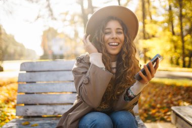 Şapkalı ve telefonlu neşeli bir kadın sonbahar parkında ahşap bir bankta oturuyor. Güzel bir kadın güneşli bir parkta rahatlar, telefonda konuşur. Hafta sonu ve eğlence konsepti.