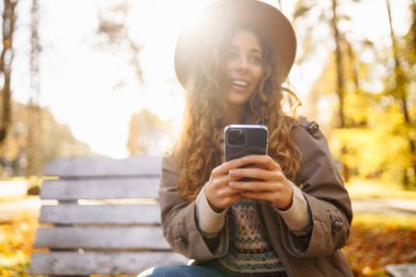 Şapkalı ve telefonlu neşeli bir kadın sonbahar parkında ahşap bir bankta oturuyor. Güzel bir kadın güneşli bir parkta rahatlar, telefonda konuşur. Hafta sonu ve eğlence konsepti.