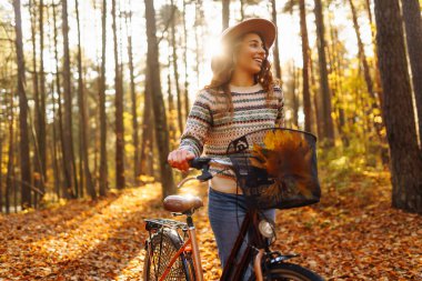 Parlak sonbahar ormanında rahat süveter ve şapka giyen bisiklet süren genç bir kadın. Neşeli bir kadın bisiklet sürüyor ve sonbahar yapraklarıyla kaplı yolda yürüyor. Etkin yaşam biçimi.