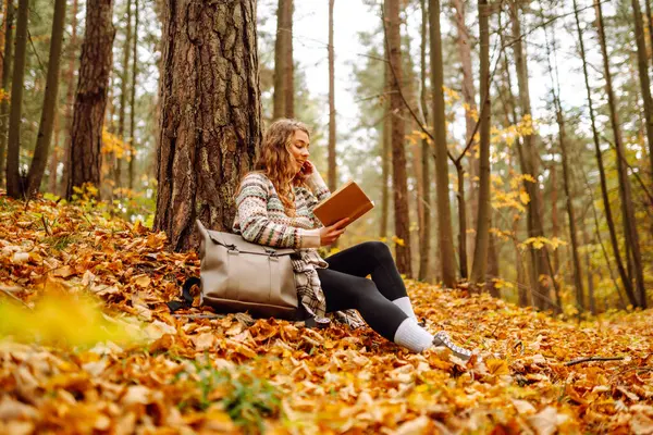 Elinde kitap olan rahat bir kadın sonbahar parkında bir ağacın yanında oturuyor. Şapkalı ve sırt çantalı güzel bir gezgin parlak yapraklarla çevrili bir kitap okuyor. Rahatlama konsepti, mevsimlik.