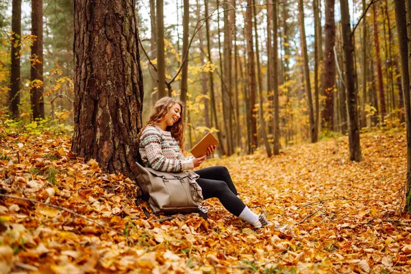 Elinde kitap olan rahat bir kadın sonbahar parkında bir ağacın yanında oturuyor. Şapkalı ve sırt çantalı güzel bir gezgin parlak yapraklarla çevrili bir kitap okuyor. Rahatlama konsepti, mevsimlik.