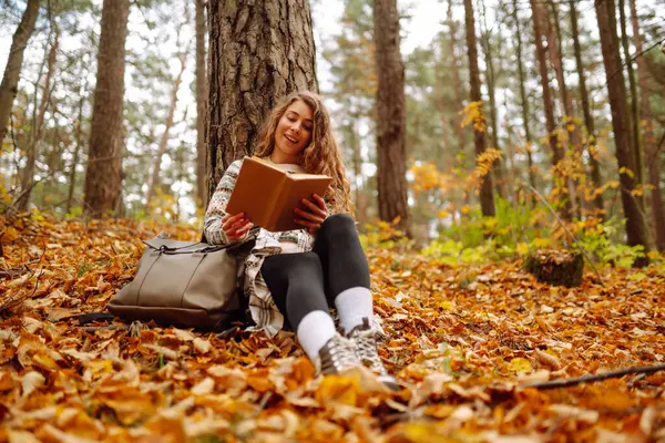 Elinde kitap olan rahat bir kadın sonbahar parkında bir ağacın yanında oturuyor. Şapkalı ve sırt çantalı güzel bir gezgin parlak yapraklarla çevrili bir kitap okuyor. Rahatlama konsepti, mevsimlik.