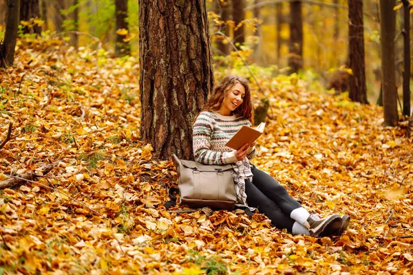 Elinde kitap olan rahat bir kadın sonbahar parkında bir ağacın yanında oturuyor. Şapkalı ve sırt çantalı güzel bir gezgin parlak yapraklarla çevrili bir kitap okuyor. Rahatlama konsepti, mevsimlik.