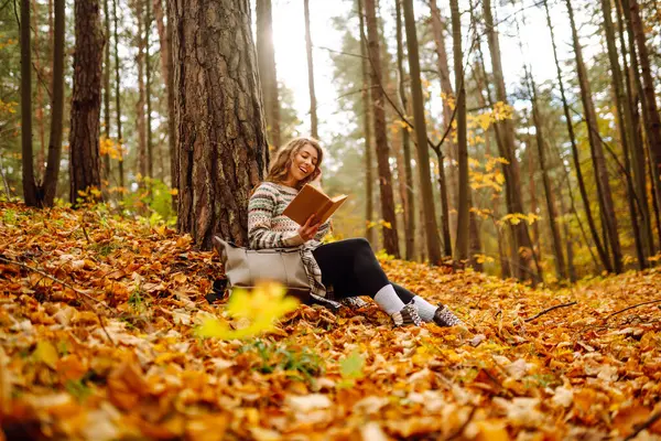 Elinde kitap olan rahat bir kadın sonbahar parkında bir ağacın yanında oturuyor. Şapkalı ve sırt çantalı güzel bir gezgin parlak yapraklarla çevrili bir kitap okuyor. Rahatlama konsepti, mevsimlik.