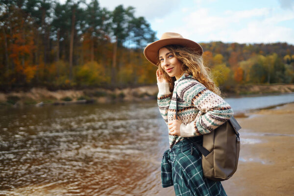 Счастливая женщина с рюкзаком у реки наслаждается солнечным днем. Женщина-путешественница чувствует себя свободной и весело на воде в осеннем лесу. Концепция приключений и развлечений.