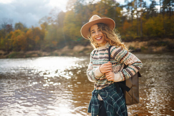 Счастливая женщина с рюкзаком у реки наслаждается солнечным днем. Женщина-путешественница чувствует себя свободной и весело на воде в осеннем лесу. Концепция приключений и развлечений.