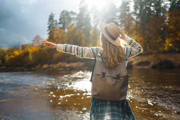 Счастливая женщина с рюкзаком у реки наслаждается солнечным днем. Женщина-путешественница чувствует себя свободной и весело на воде в осеннем лесу. Концепция приключений и развлечений.