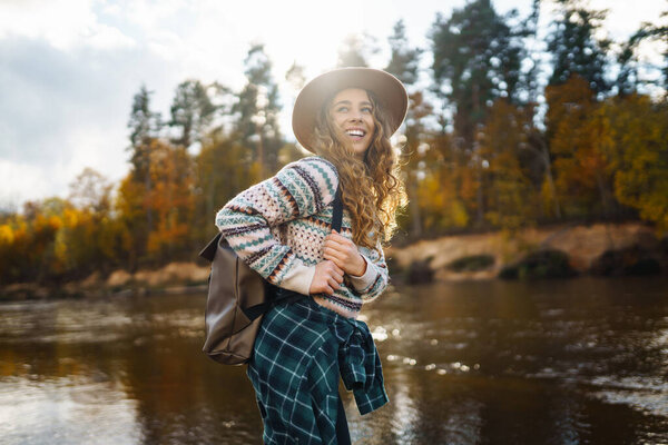 Счастливая женщина с рюкзаком у реки наслаждается солнечным днем. Женщина-путешественница чувствует себя свободной и весело на воде в осеннем лесу. Концепция приключений и развлечений.