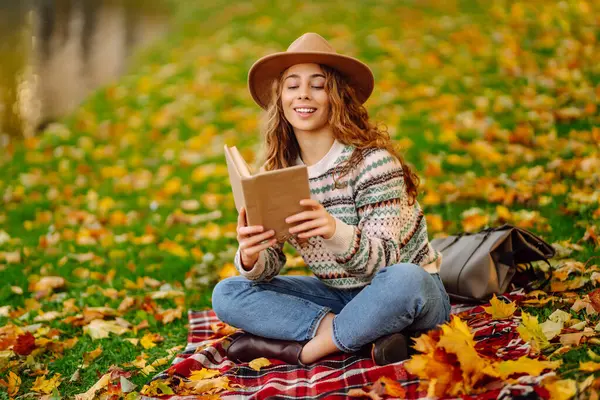 Sonbahar parkında kırmızı battaniyede oturan şapkalı genç bir kadın. Kitap okumayı seven mutlu bir kadın ve sarı yapraklı çayırda güneşli bir gün. Dinlenme kavramı, rahatlama..