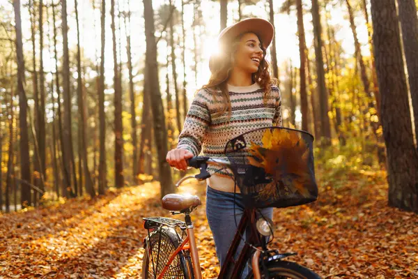 Parlak sonbahar ormanında rahat süveter ve şapka giyen bisiklet süren genç bir kadın. Neşeli bir kadın bisiklet sürüyor ve sonbahar yapraklarıyla kaplı yolda yürüyor. Etkin yaşam biçimi.