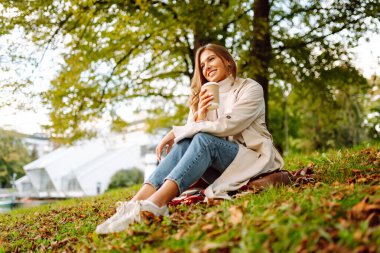 Sonbahar parkında bir bardak sıcak içecekle battaniyede oturan yağmurluk giymiş gülümseyen kadın. Güzel bir kadın göl kenarında eğleniyor. Yürüyüş kavramı, dinlenme.