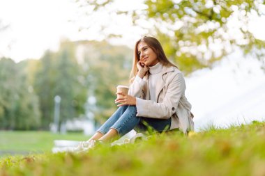 Sonbahar parkında bir bardak sıcak içecekle battaniyede oturan yağmurluk giymiş gülümseyen kadın. Güzel bir kadın göl kenarında eğleniyor. Yürüyüş kavramı, dinlenme.